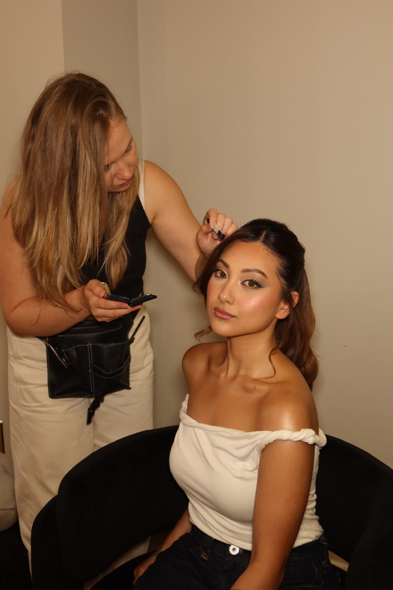A woman with long dark hair sits on a black chair, wearing an off-shoulder white top, with a makeup artist working on her hair.