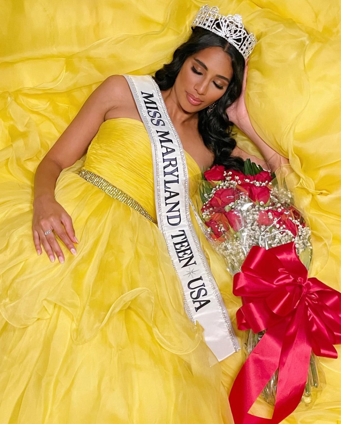 Woman wearing a crown and sash, lying on yellow fabric, holding a bouquet with a large red ribbon, smiling.