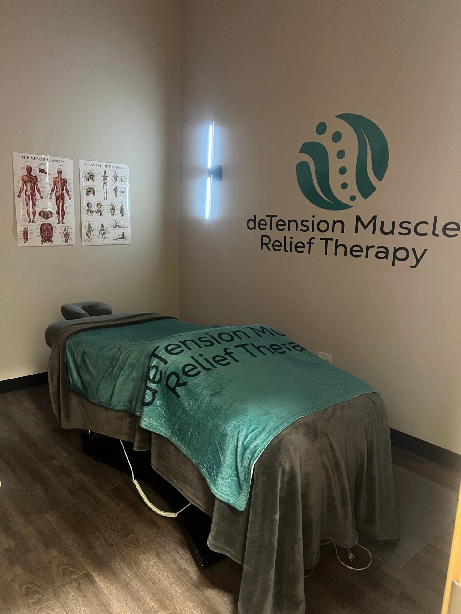 A massage table covered with a teal sheet in a therapy room with a wall sign and anatomical posters.