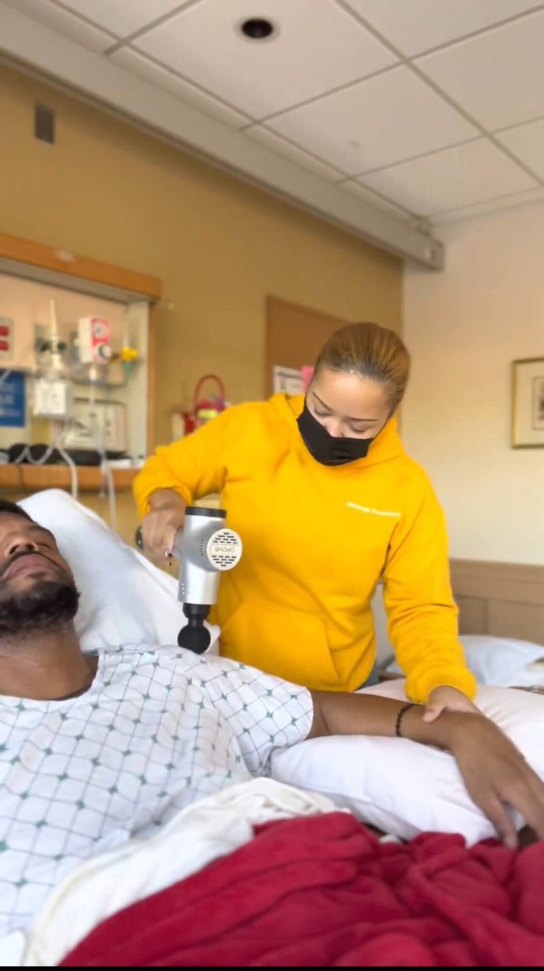 Healthcare worker in yellow uniform attends to a patient in hospital bed, wearing a face mask, in a clinical setting.