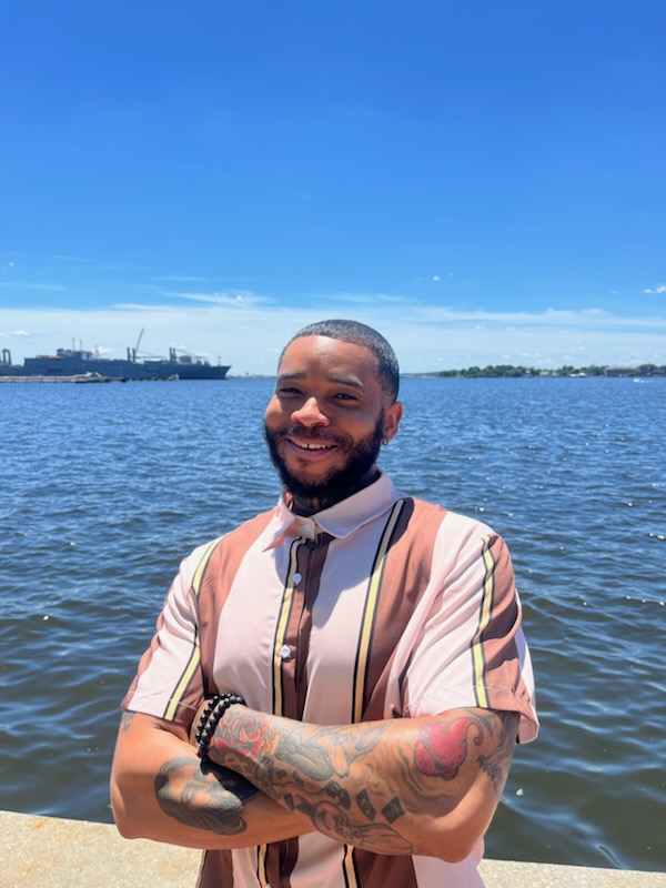 Man with a beard and tattoos smiling outdoors near water, with a cityscape in the background.