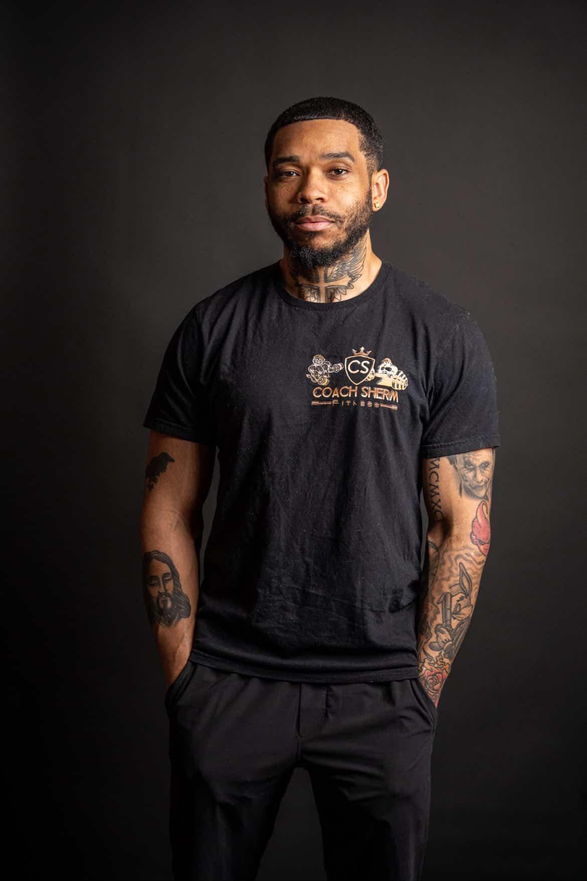 Man with tattoos on arms and beard, wearing a black T-shirt with a logo, standing against a dark background.