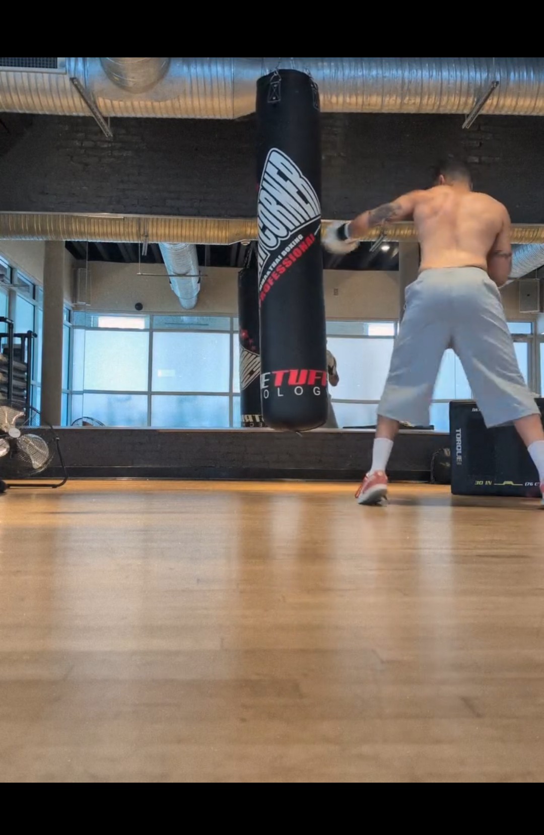 Man in gray shorts and no shirt standing in a gym with punching bag and equipment, viewed from low angle.