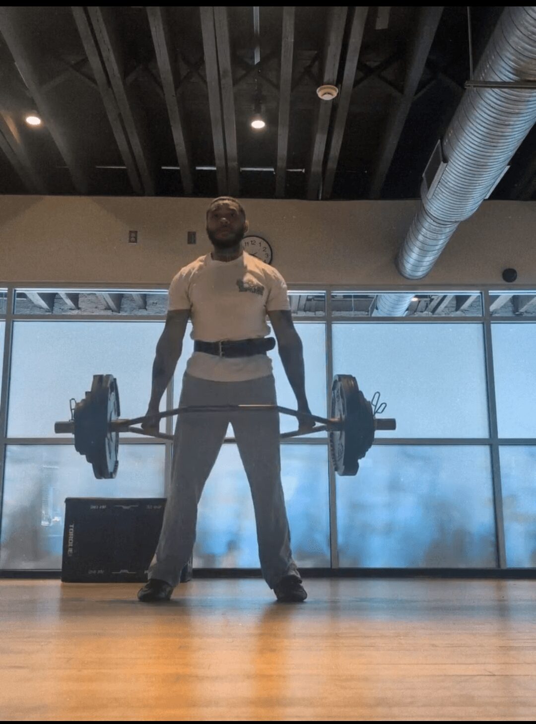 Person lifting a barbell in a gym with large windows and ceiling pipes visible.