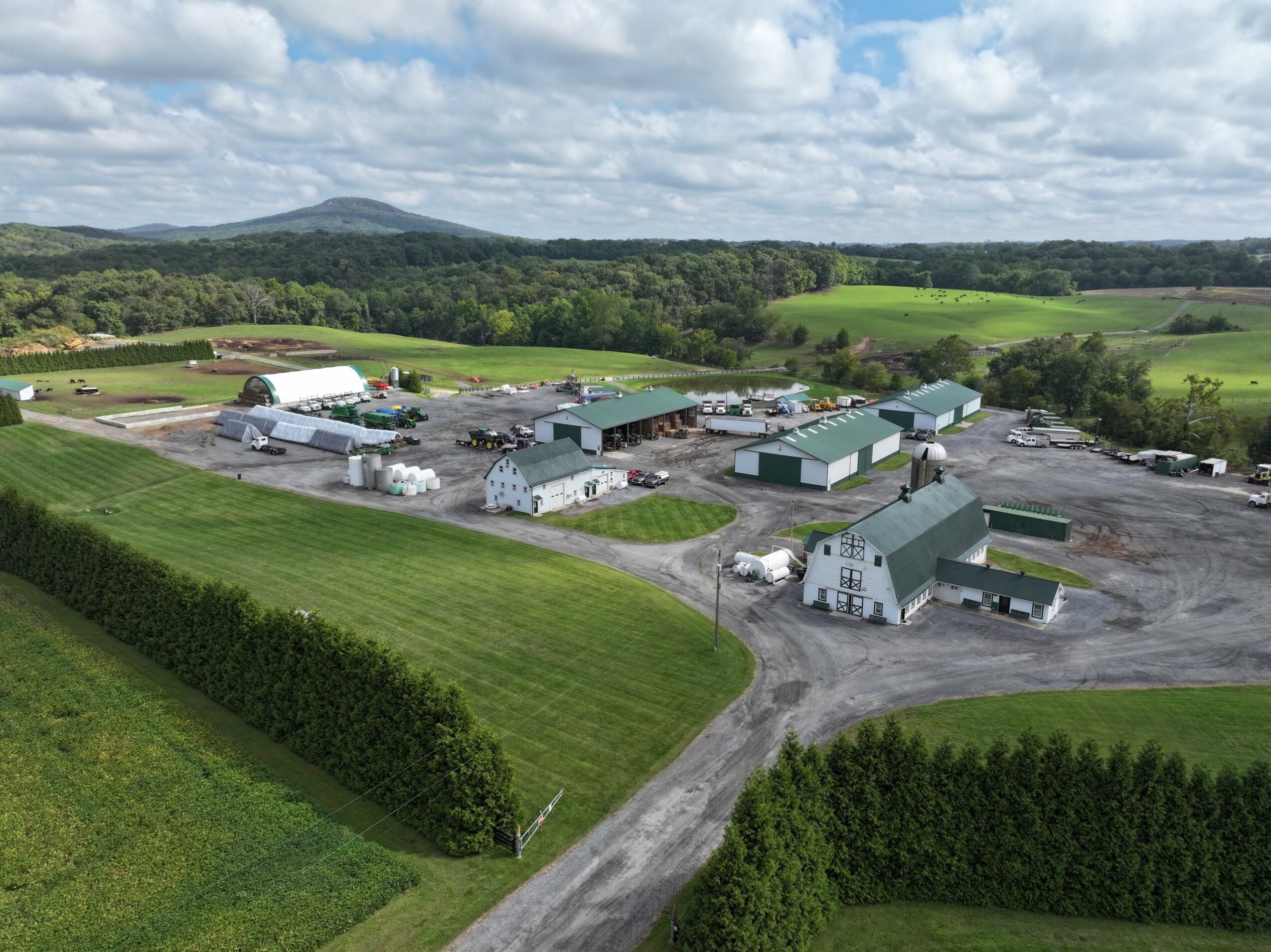 Aerial view of a farm with multiple buildings, surrounded by green fields and hills under a partly cloudy sky.