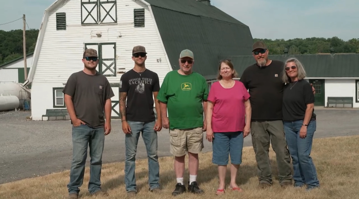 Group of six people standing outdoors in front of a white barn, smiling, with a grassy area and trees in the background.