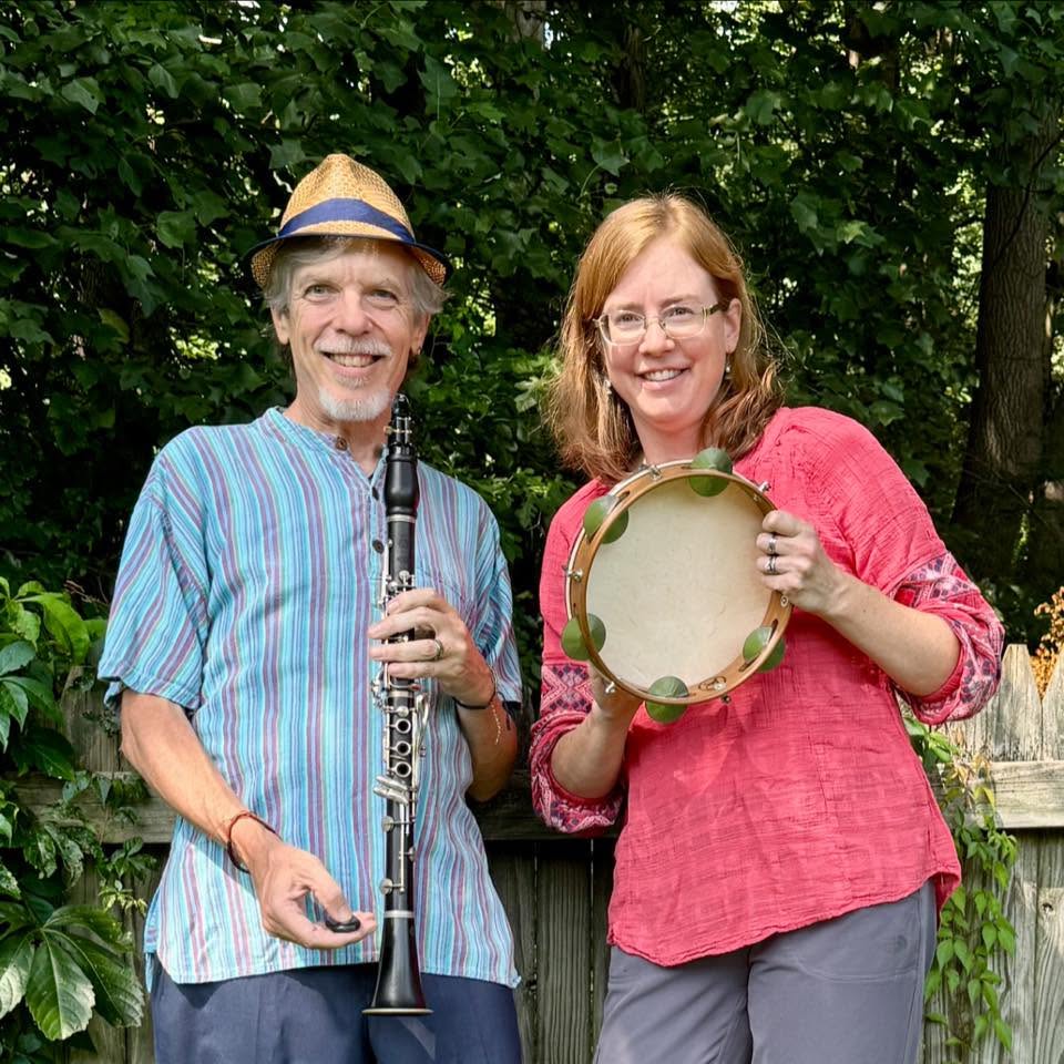 Two people smiling outdoors, one holding a clarinet and the other holding a tambourine, standing in front of green foliage.