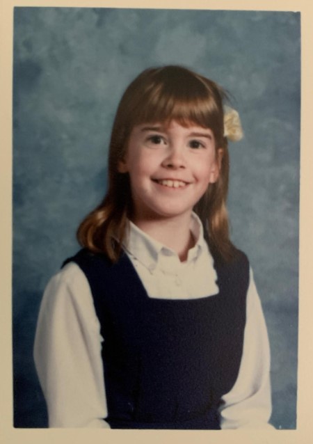 Young girl with shoulder-length hair, smiling, wearing a white shirt and dark vest, against a blue background.