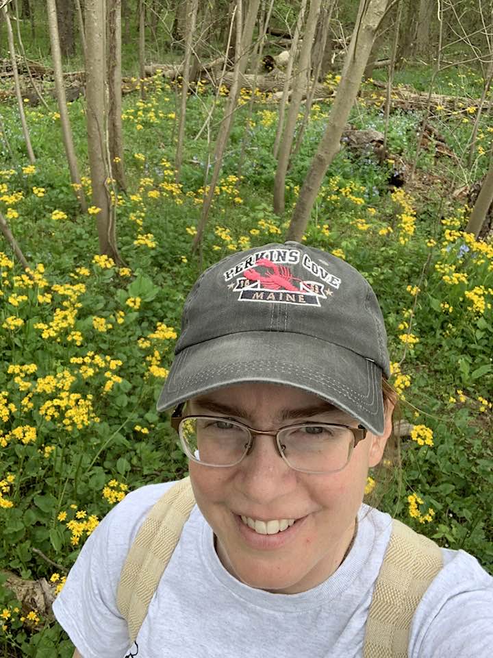 Person wearing a cap and glasses smiling outdoors among yellow flowers and trees.