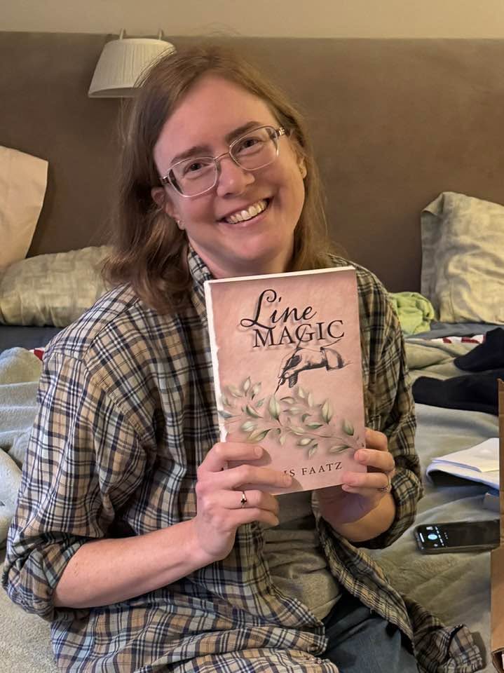 Person with glasses smiling holding a book titled 'Line Magic' in a room with a bed and pillows.