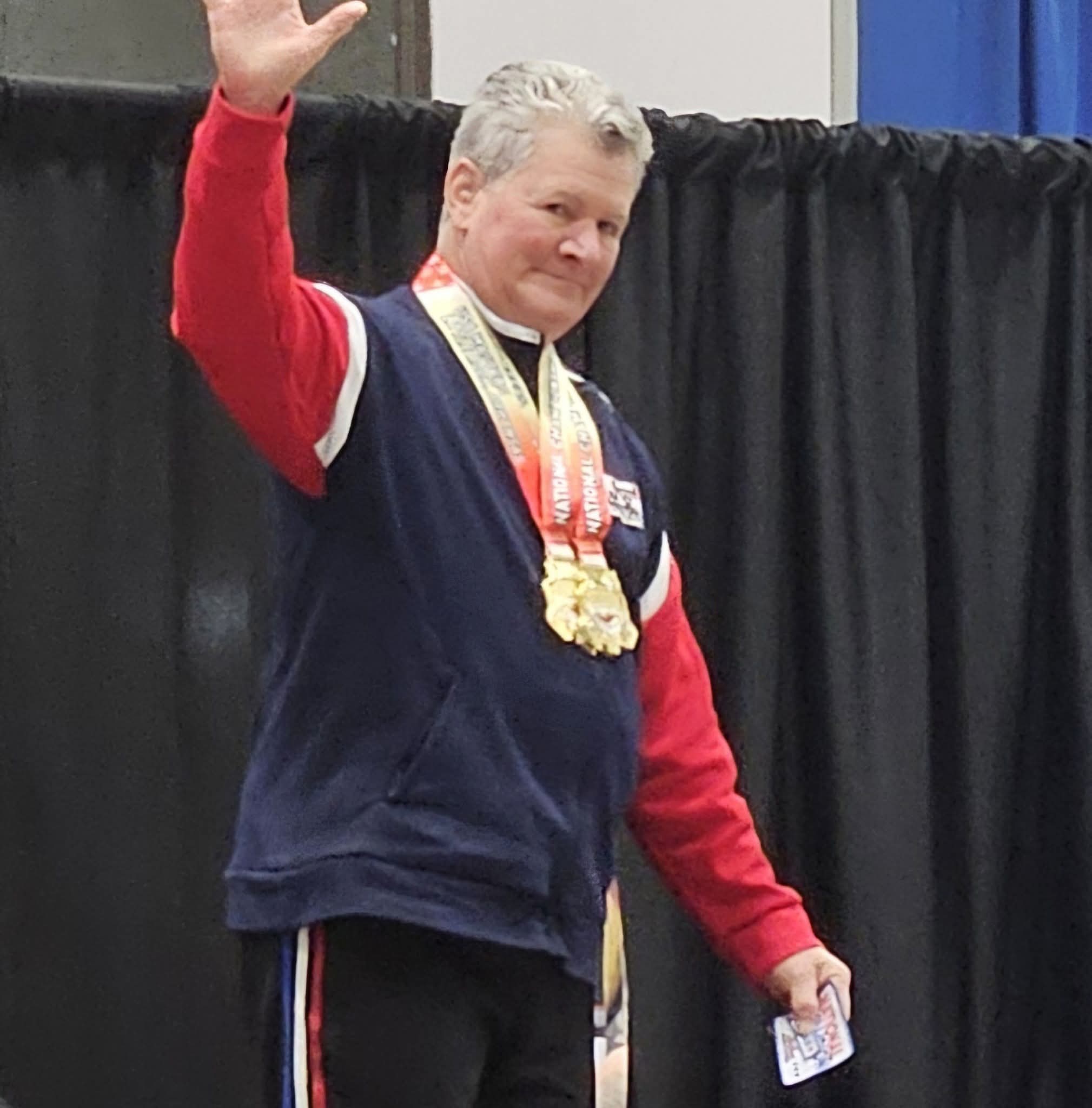Woman with medals around neck waving, standing in front of black curtain, wearing a navy and red jacket, holding a paper.