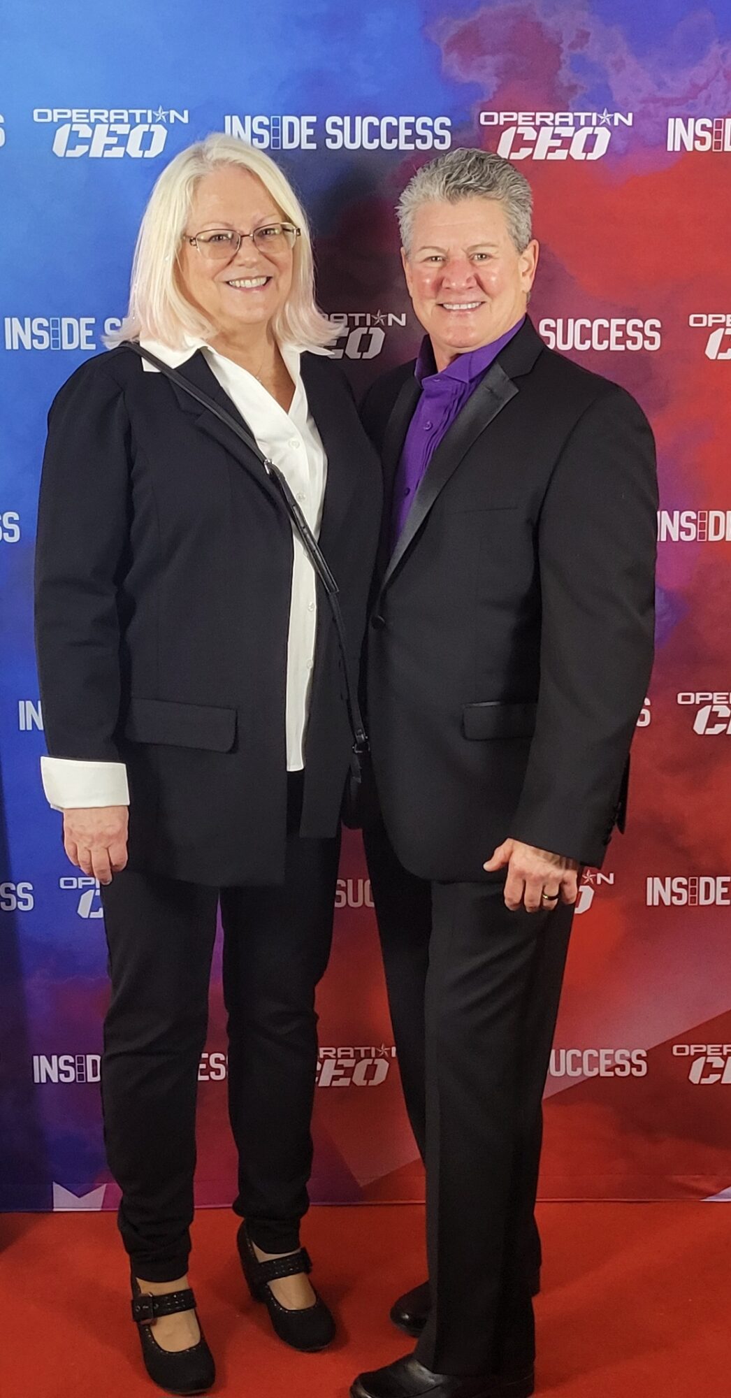 Two people, a woman and a man, stand together smiling in front of a red and blue backdrop with text.