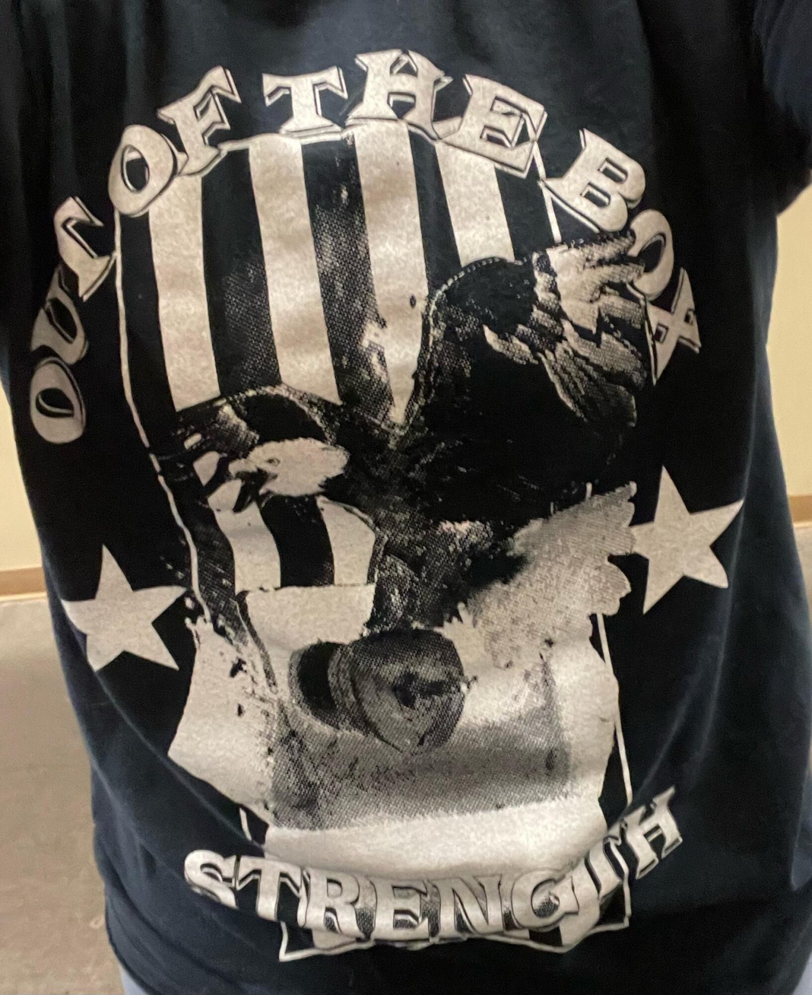 Person wearing a black T-shirt with a graphic of a dog, stars, and text reading 'Out of the Box Strength'.