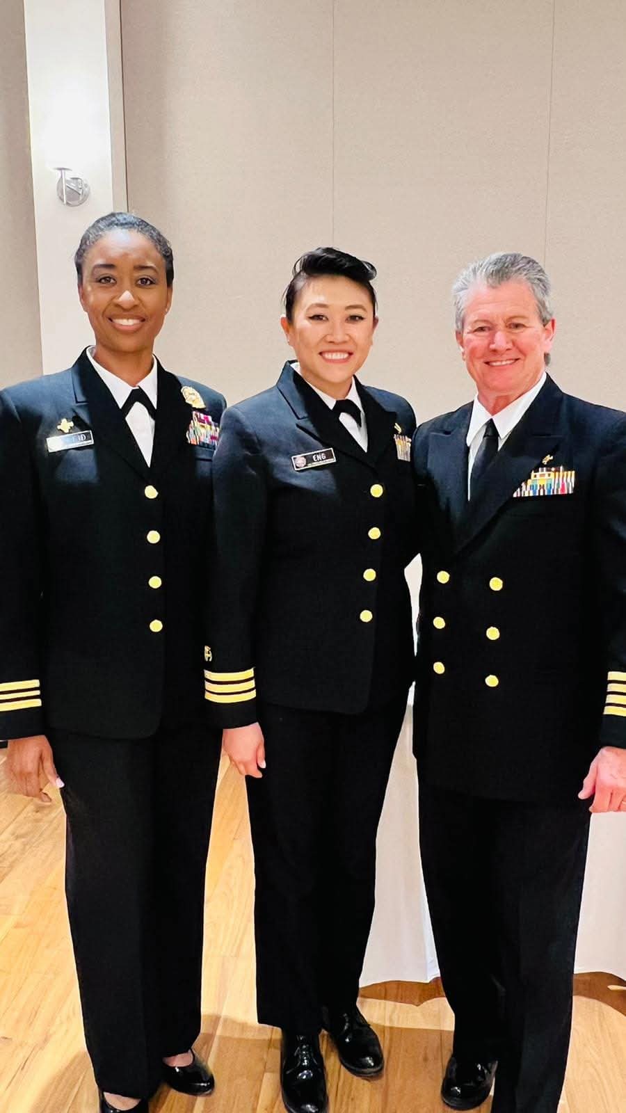 Three people in military uniforms standing together indoors, smiling, with a plain background.