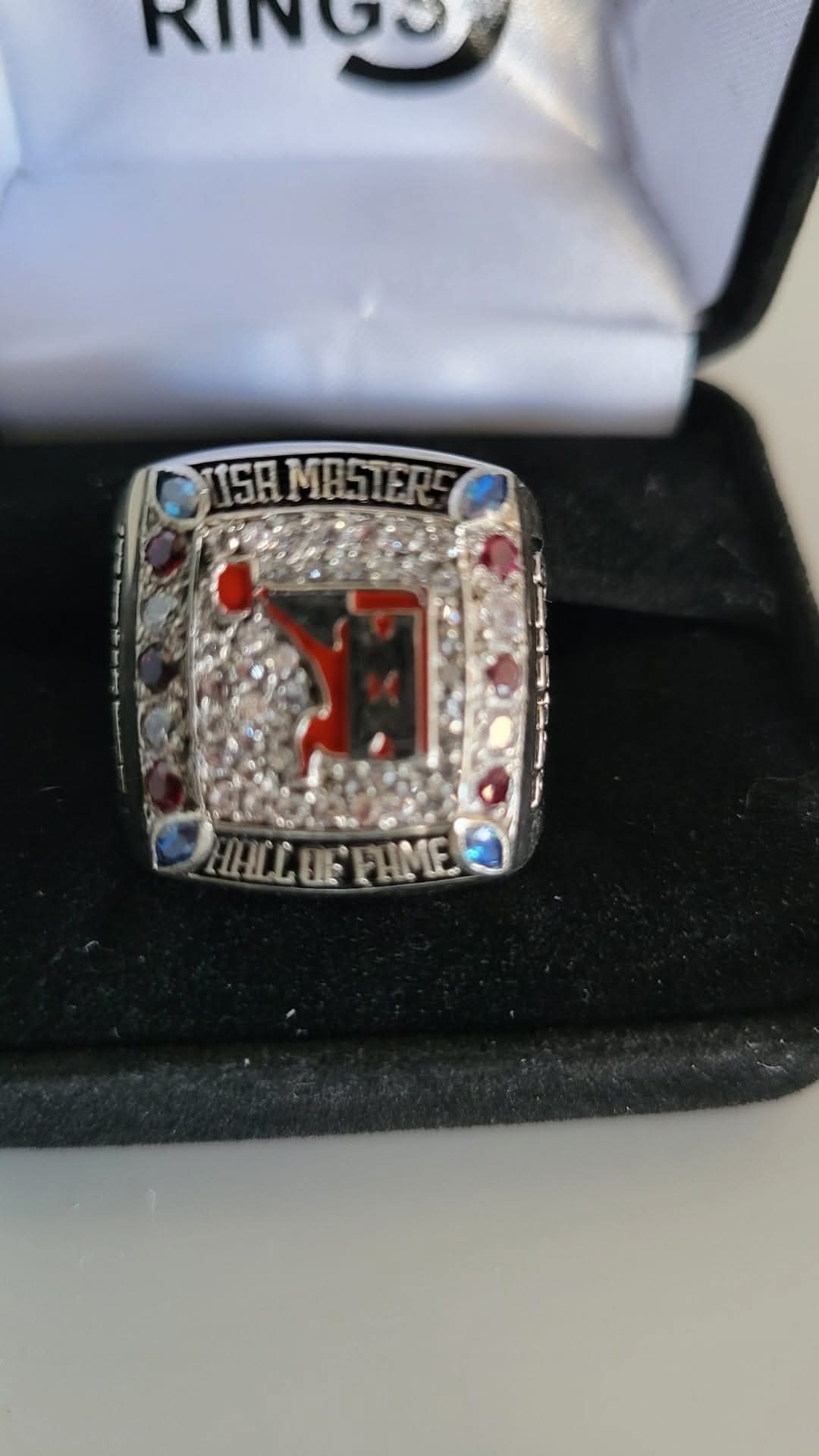 A championship ring with a central red and black emblem, surrounded by small gemstones, and engraved text, placed on a black surface.