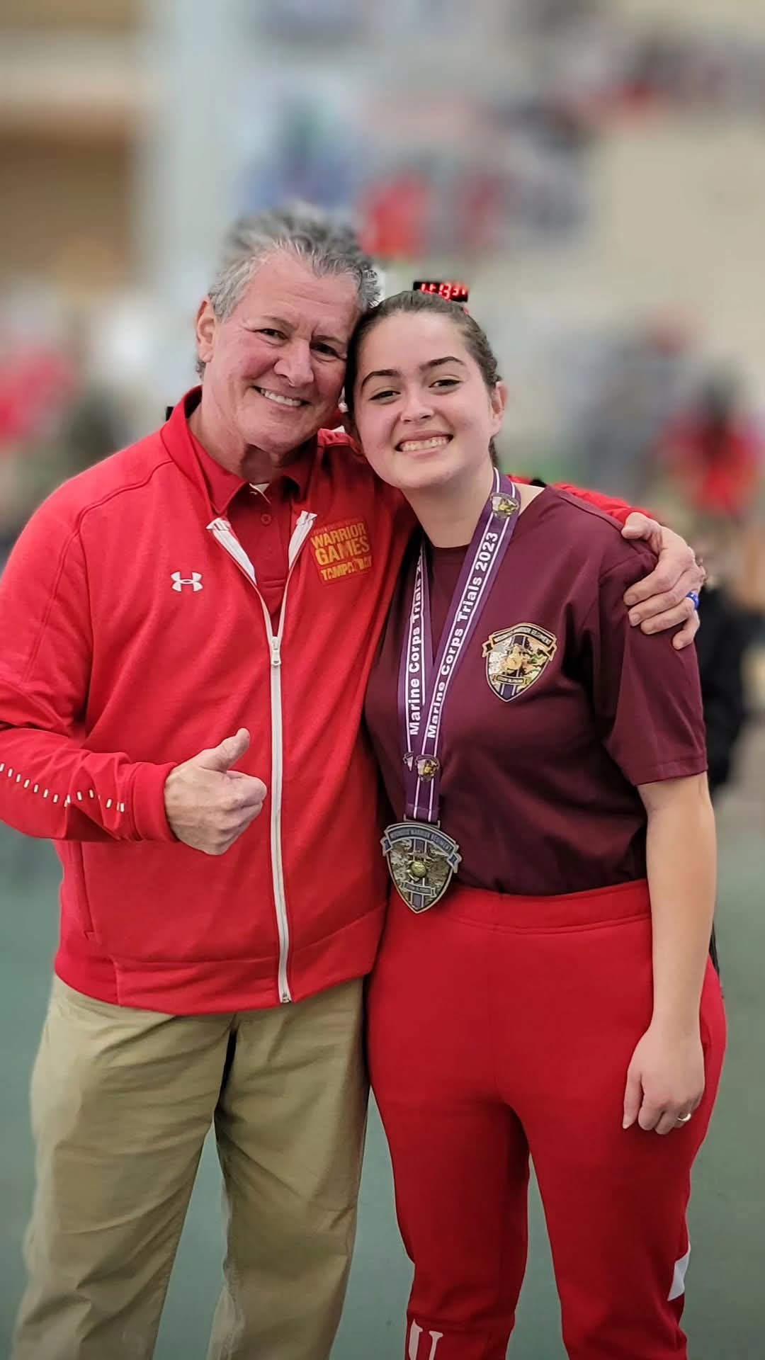 Two smiling women, one in a red jacket and the other in a maroon shirt with medals, stand outdoors with arms around each other.