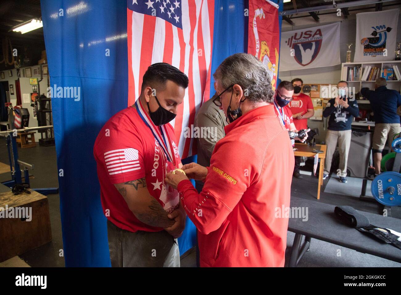 Two men in red shirts and face masks exchange medals at a ceremony with American flags in the background.