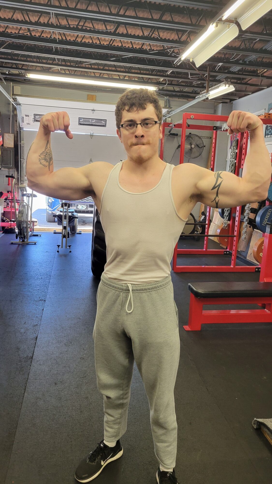 Person in gym wearing white tank top and gray sweatpants, flexing arm, standing in gym equipment area.