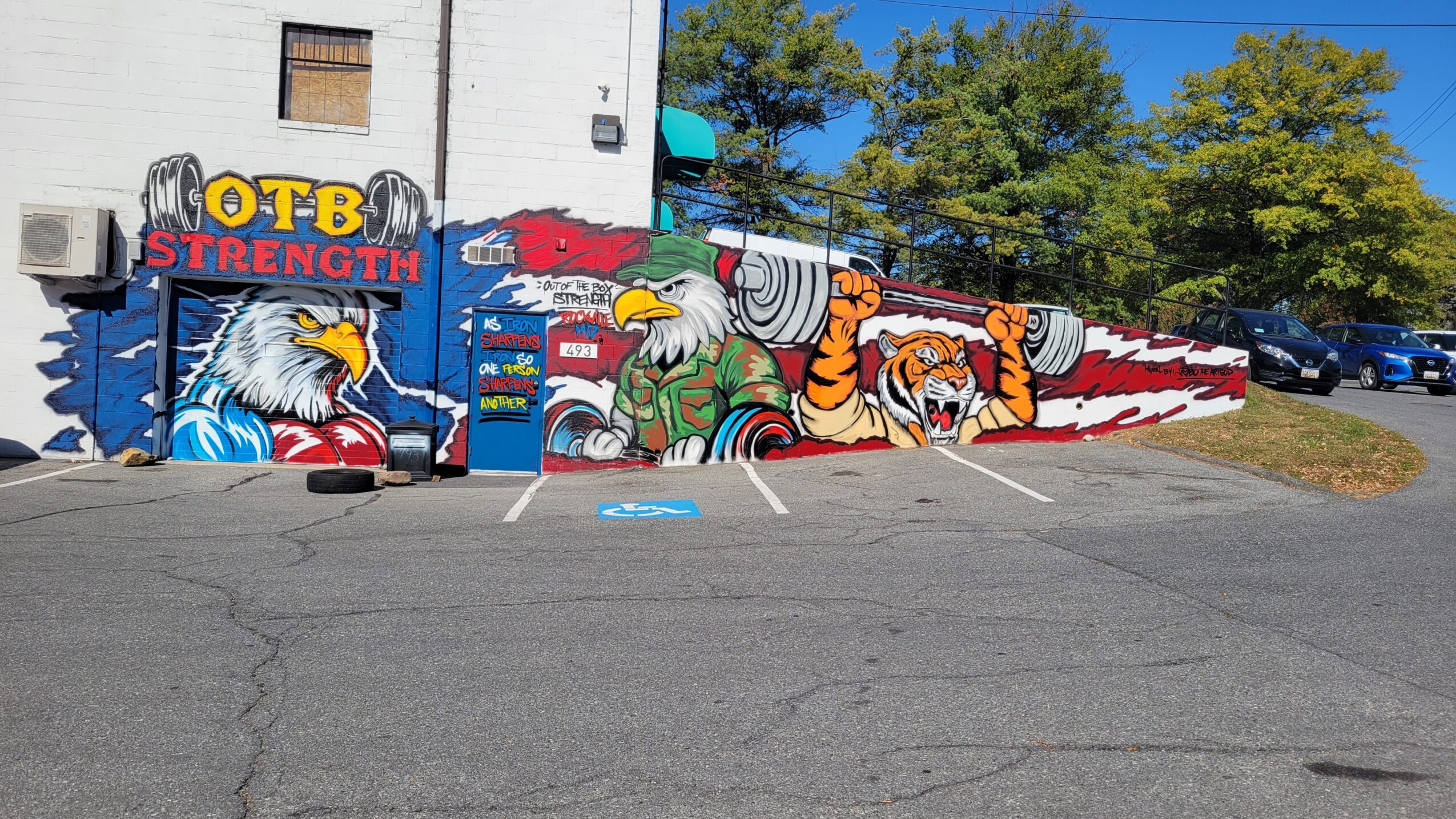 Colorful mural with eagles, tigers, and the words 'OTB Strength' on a building wall, with parking lot in foreground.