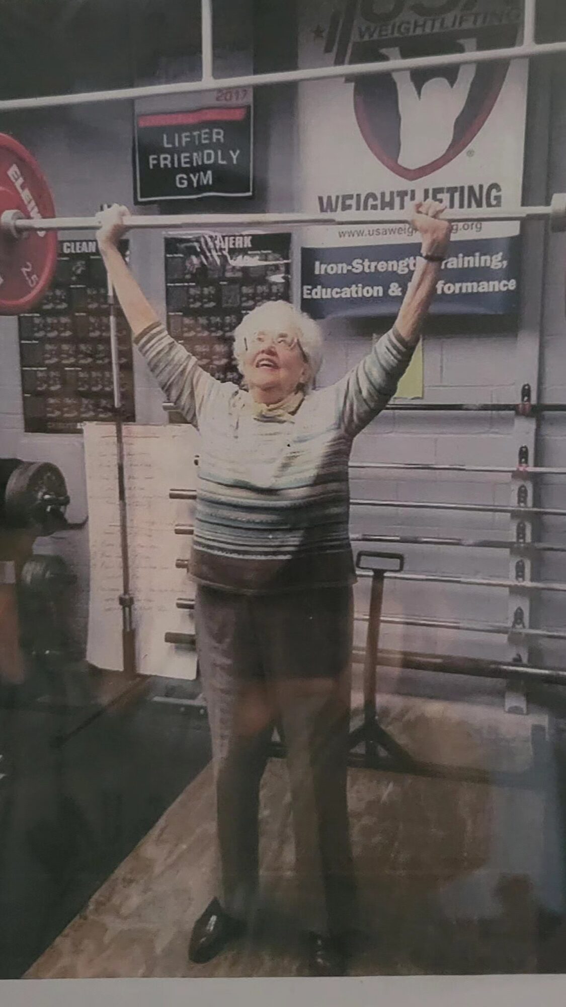 Person lifting a barbell overhead in a gym, smiling, with gym equipment and signs in the background.