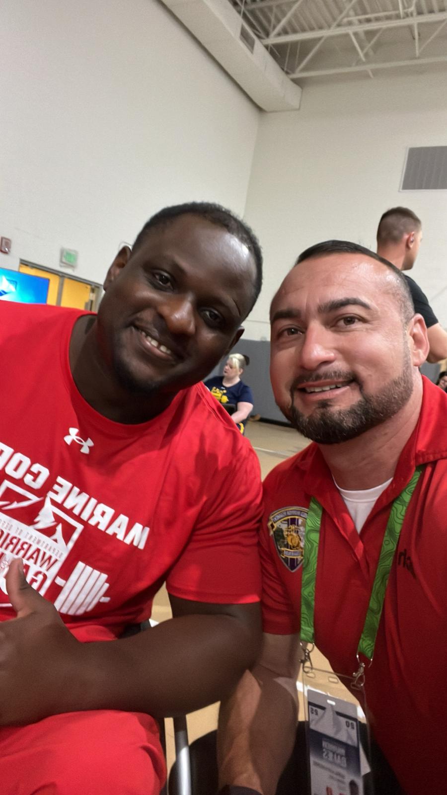 Two men smiling and taking a selfie indoors, one wearing a red sports shirt and the other a red polo shirt with a badge.