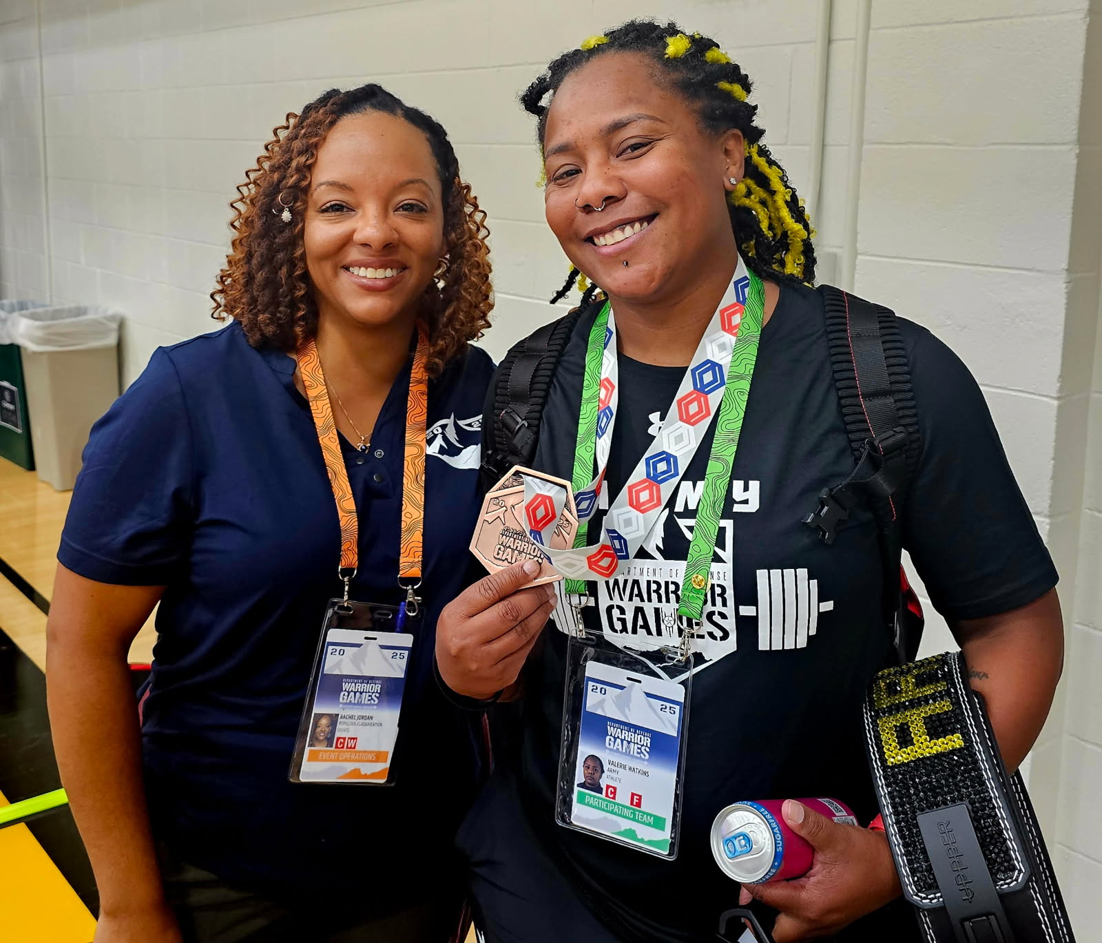 Two women smiling, one holding a medal and a water bottle, in a room with a white wall.