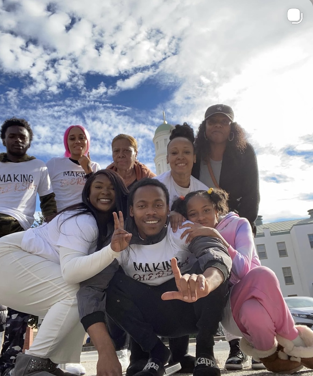 Group of seven people outdoors, smiling, some making peace signs, with a blue sky and clouds in the background.