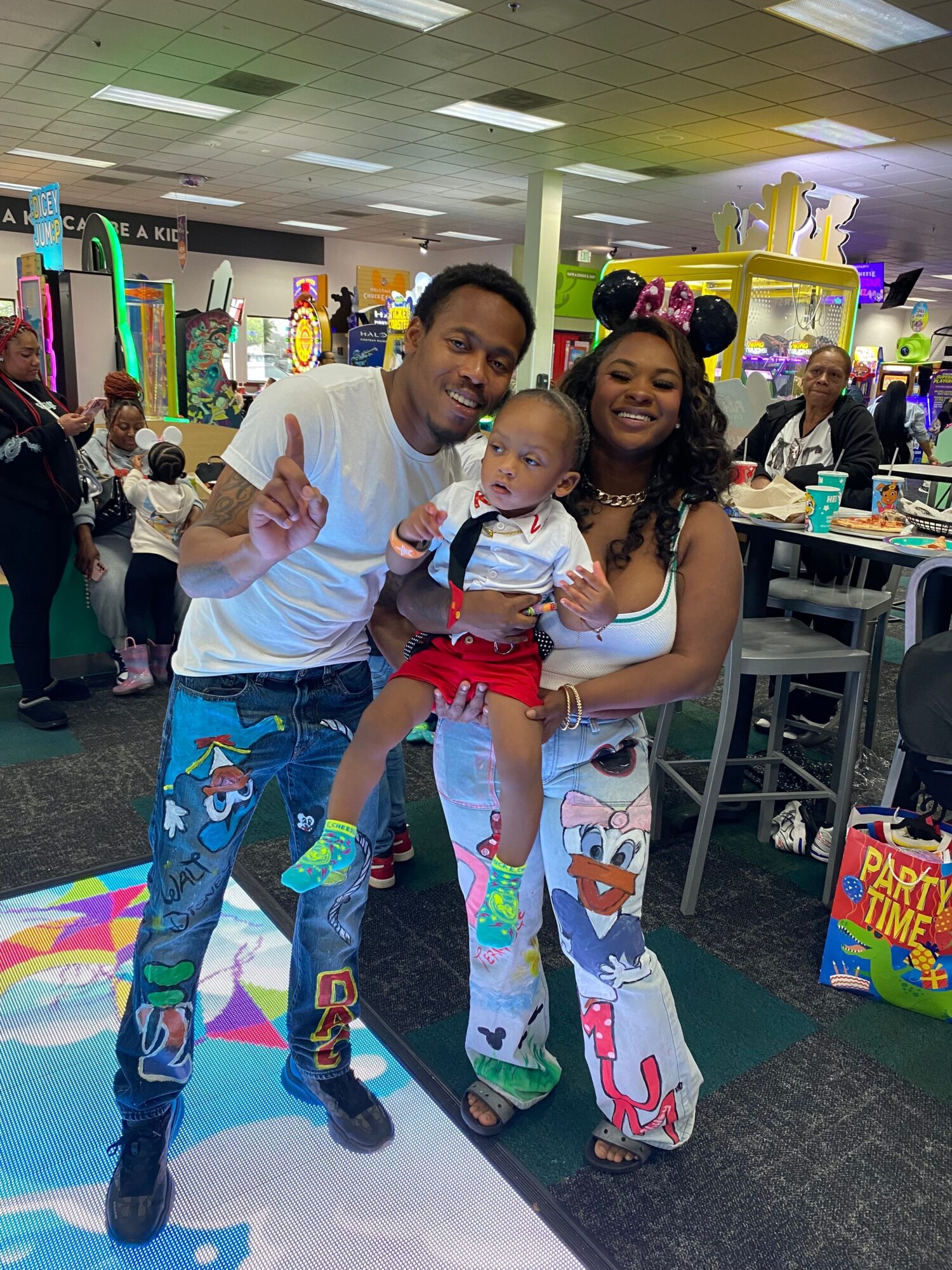 Family with two children in a shopping mall, woman holding a baby, man making peace sign, colorful background.