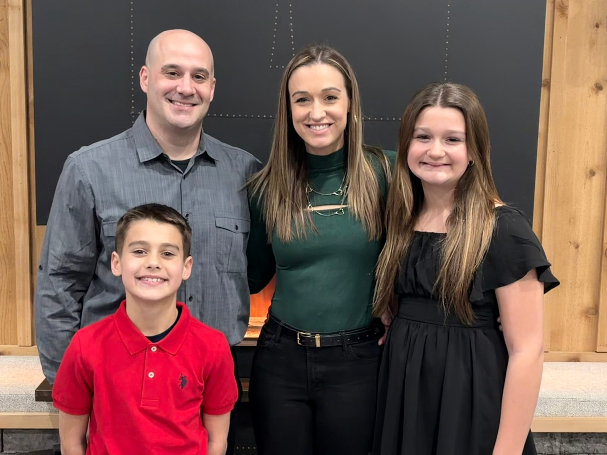 Four people, two adults and two children, smiling and standing together indoors against a dark background.