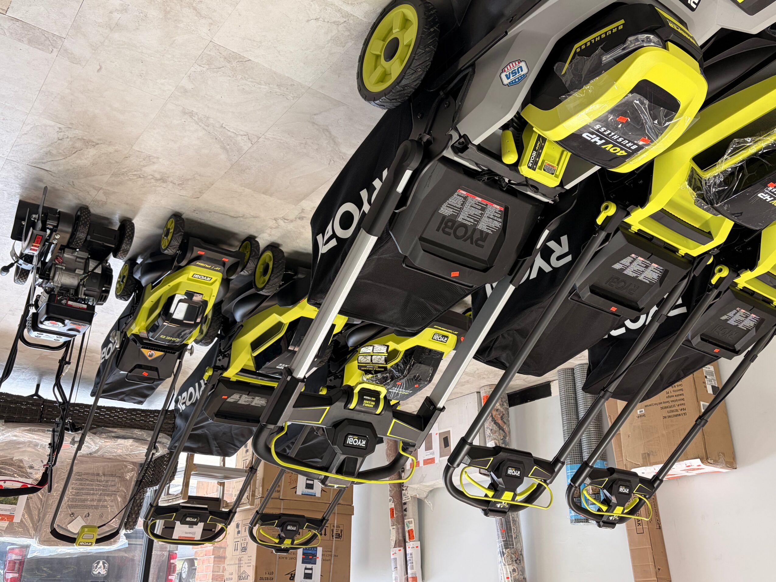 Lawnmowers and power tools arranged on a concrete floor, with some stacked and others lined up.