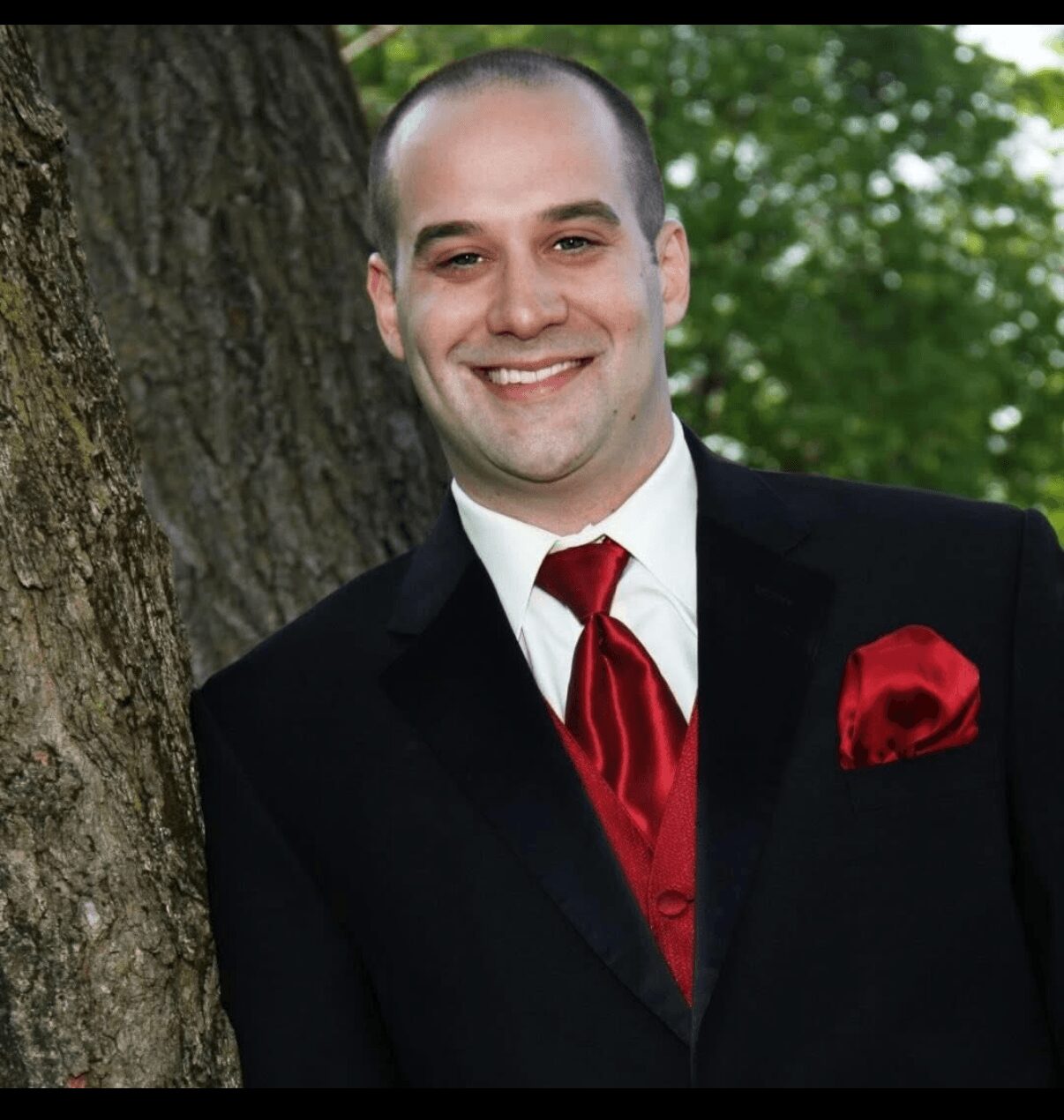 Man in formal suit with red tie and pocket square leaning against a tree outdoors, smiling, green foliage in background.