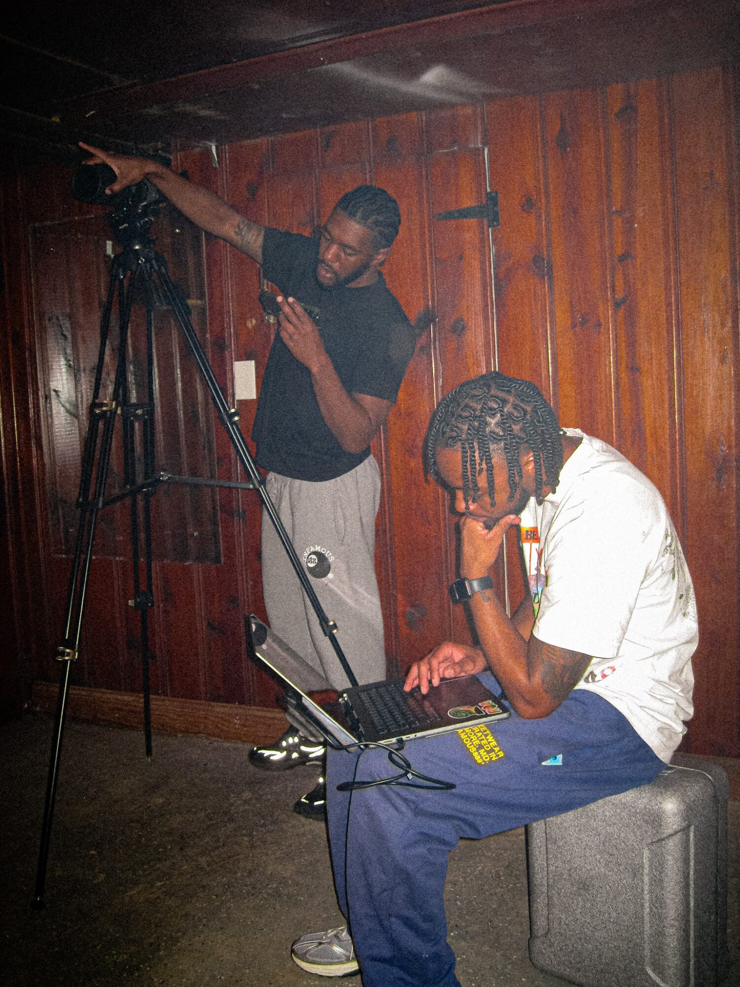 Two men in a wooden room, one adjusting a camera on a tripod, the other sitting with headphones and a laptop.