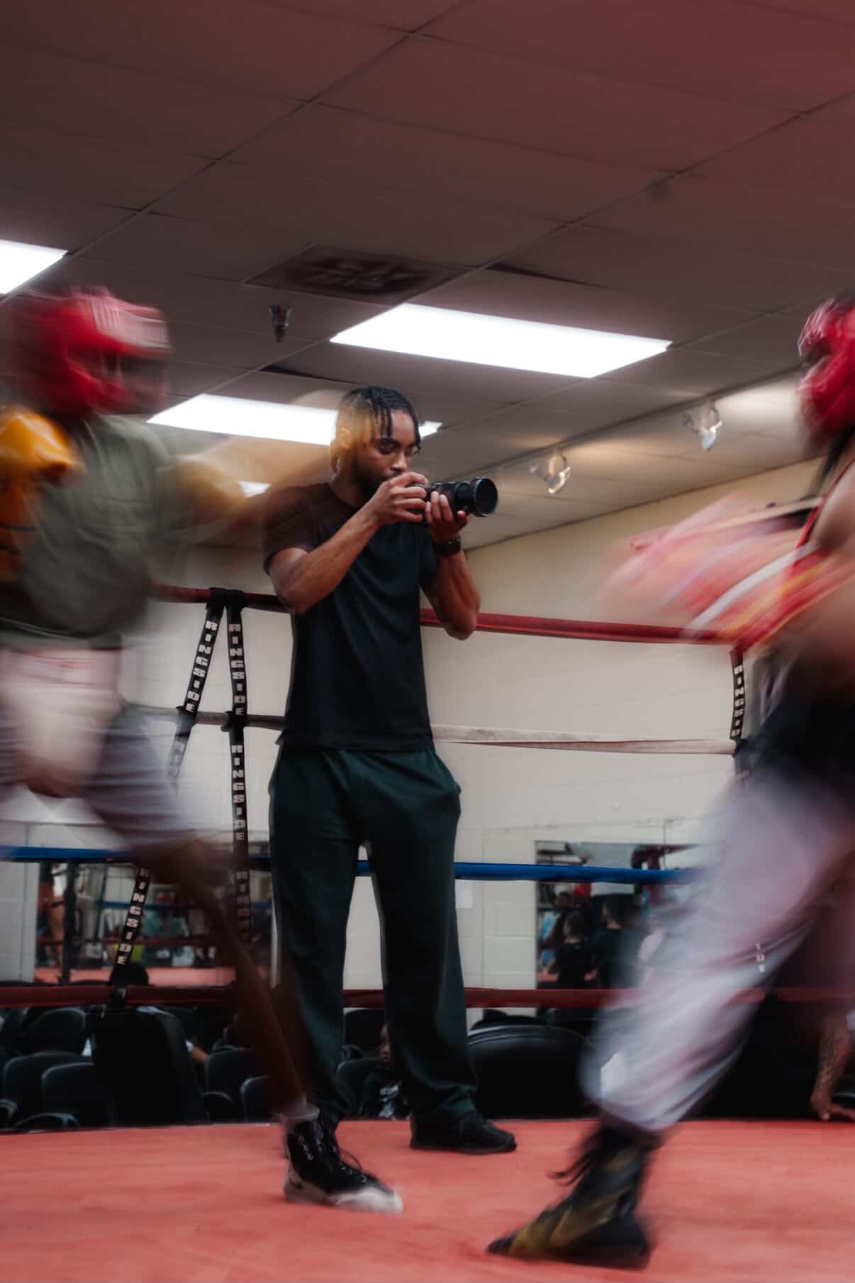 Person in black holding a camera in a boxing gym with two fighters in red headgear and gloves, blurred motion.