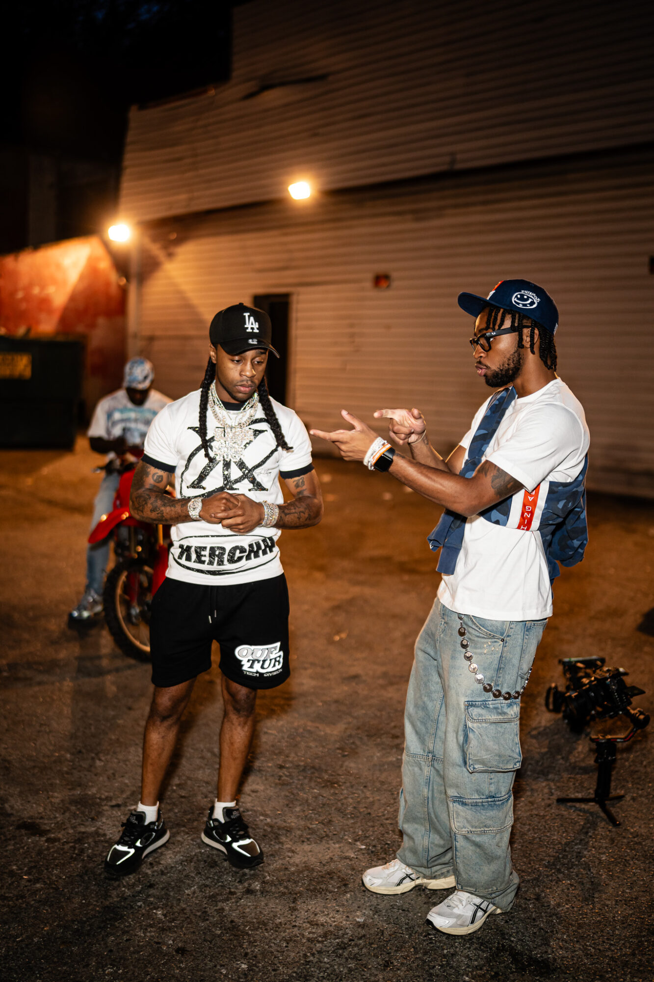 Two men stand and talk outdoors at night, one with a backpack and the other gesturing with his hands.