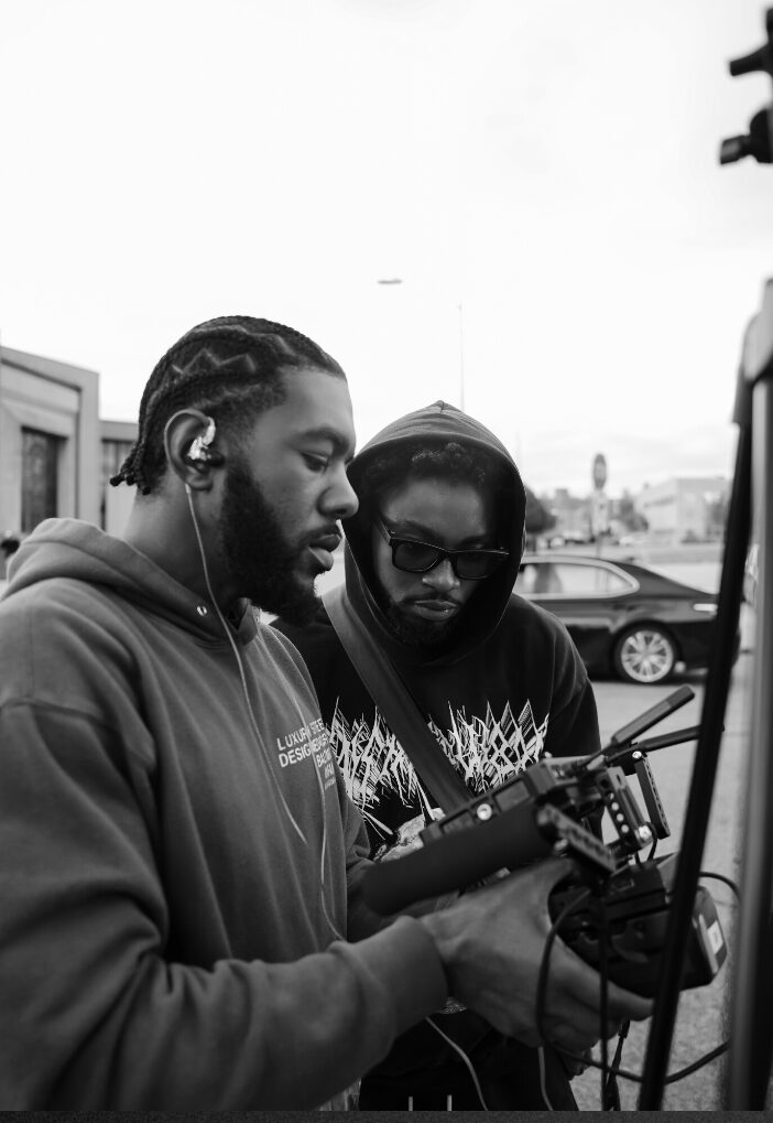 Two men with hoodies and headphones look at a device outdoors, with cars and buildings in the background.