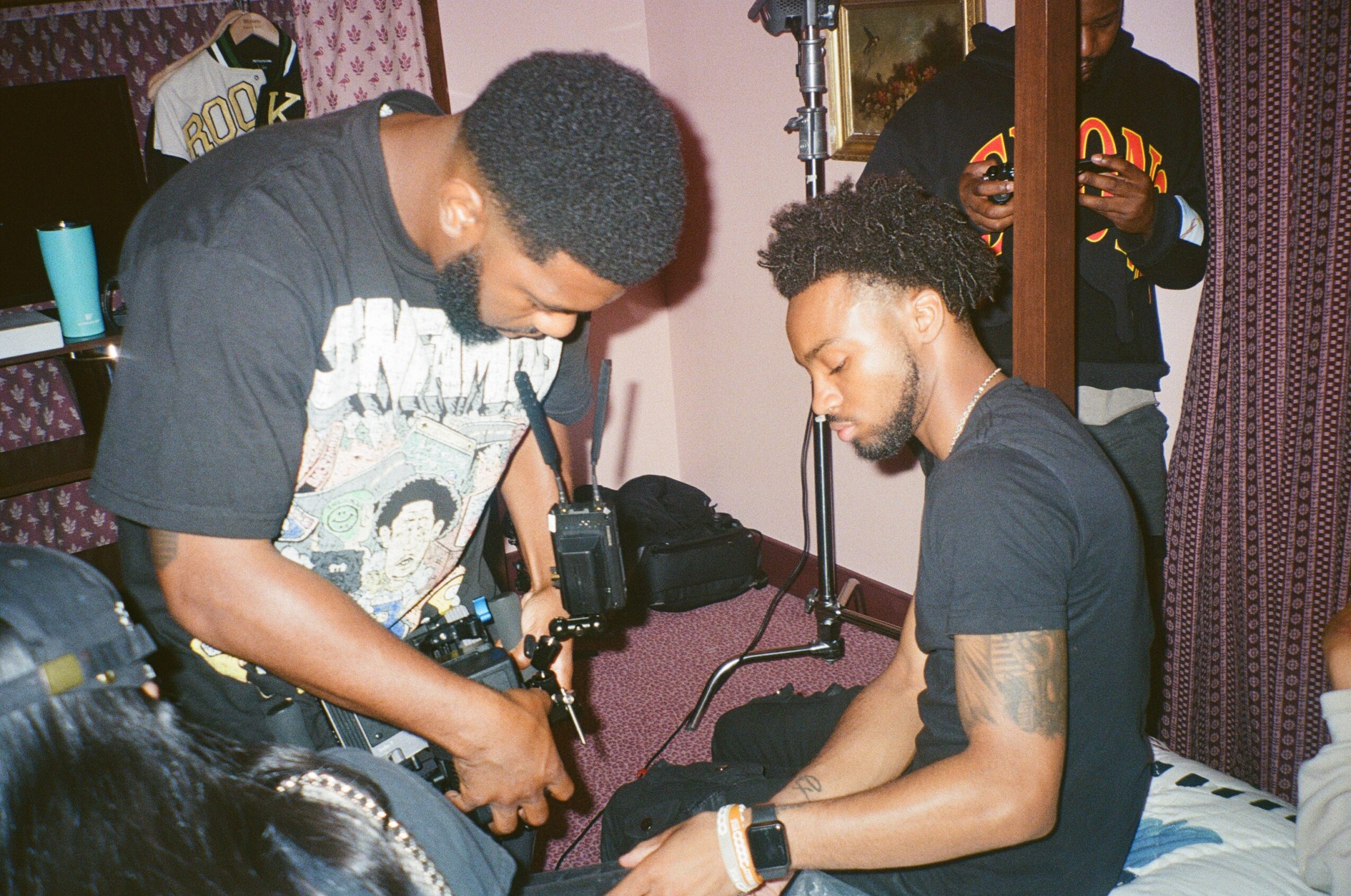 Two men are preparing for a photoshoot or filming, with one adjusting equipment and the other sitting on a bed.
