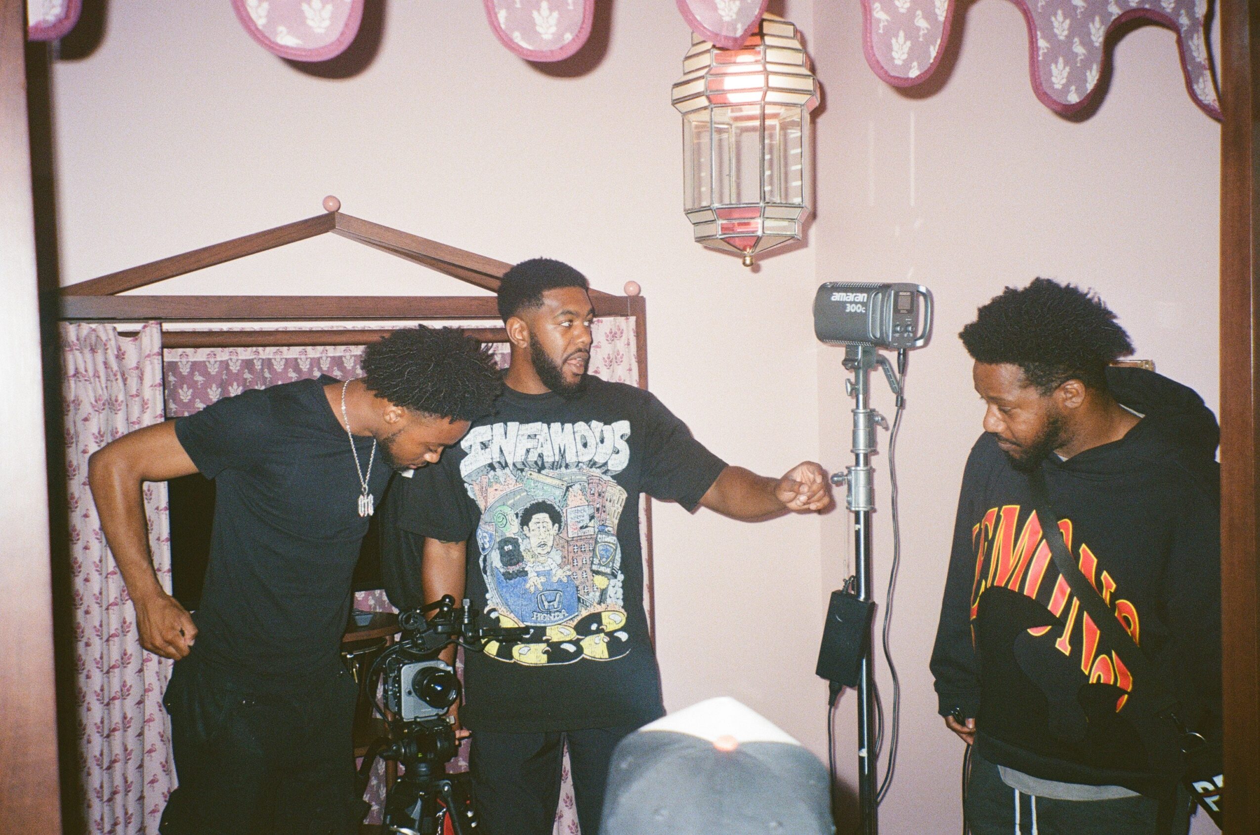 Three men in a room with pink curtains and hanging lanterns, one holding a camera, others looking at it.
