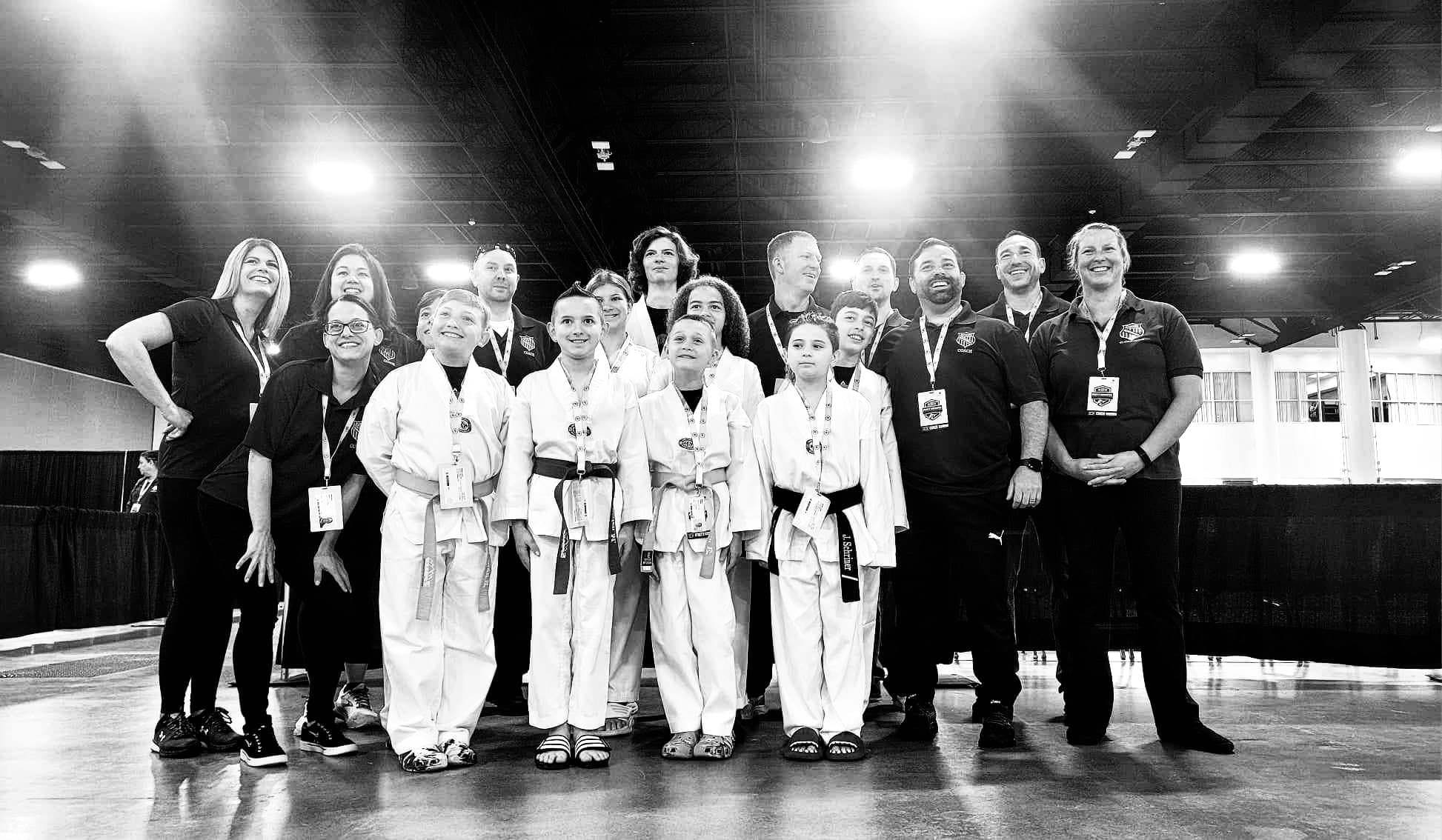 Group of children and adults posing together, some children in martial arts uniforms, in an indoor setting with bright lights.