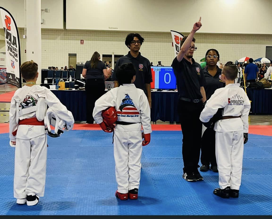 Three children in martial arts uniforms stand on a blue mat, listening to an instructor with a raised hand, in a gymnasium.
