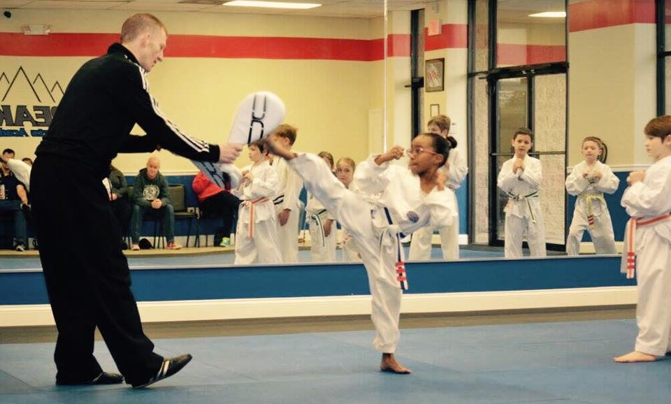 Young girl in a karate uniform performs a high kick towards an instructor holding a target pad, with children watching.