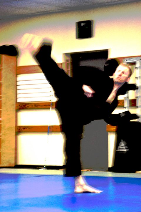 Person performing a high kick in a martial arts dojo, with a blue mat and wooden wall panels.