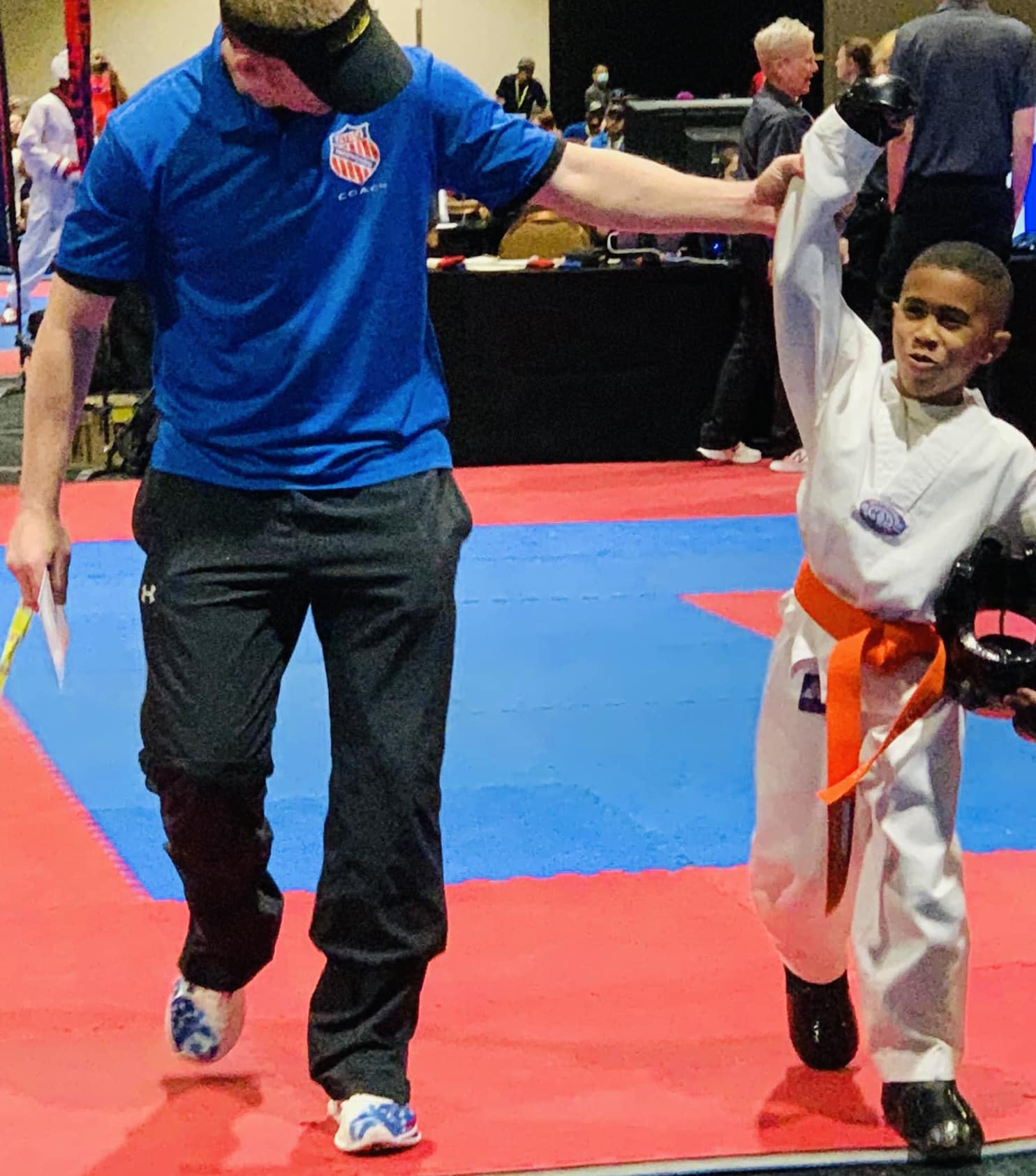 Young boy in a white martial arts uniform with an orange belt, raising his arm, next to a man in a blue shirt, on a red and blue mat.