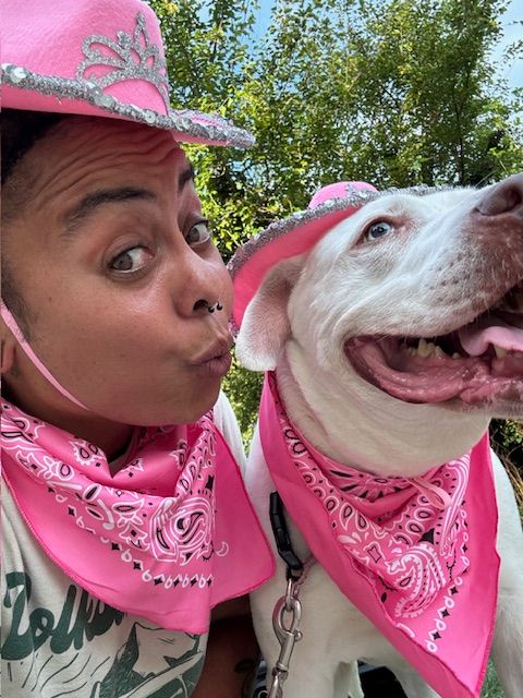 Person with a pink bandana and a pink scarf, smiling with a white dog wearing a pink bandana outdoors.