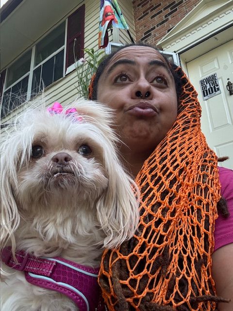 Person with dark hair and orange scarf holding a small light-colored dog with a pink collar outside a house.