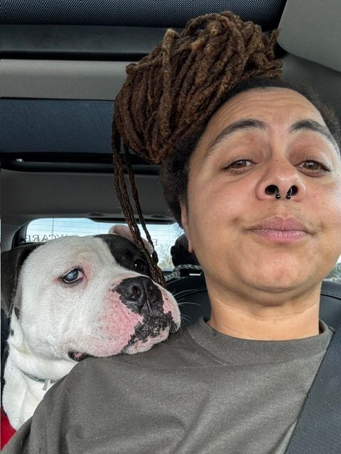 Person with dreadlocks and a dog with black spots on white fur, both looking at the camera.