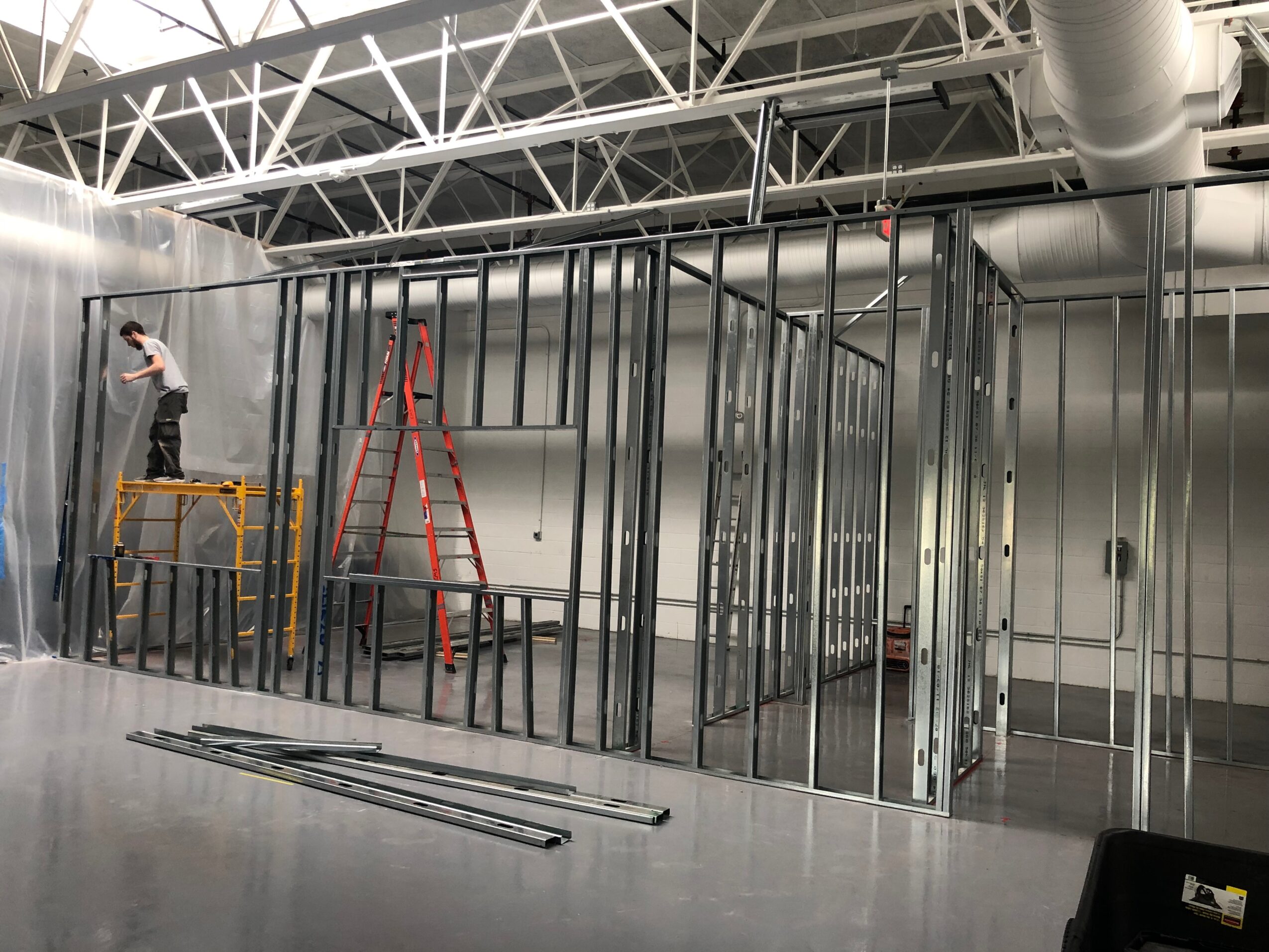 Construction site with metal framing, a worker on a yellow lift, and a red ladder inside a large industrial space.