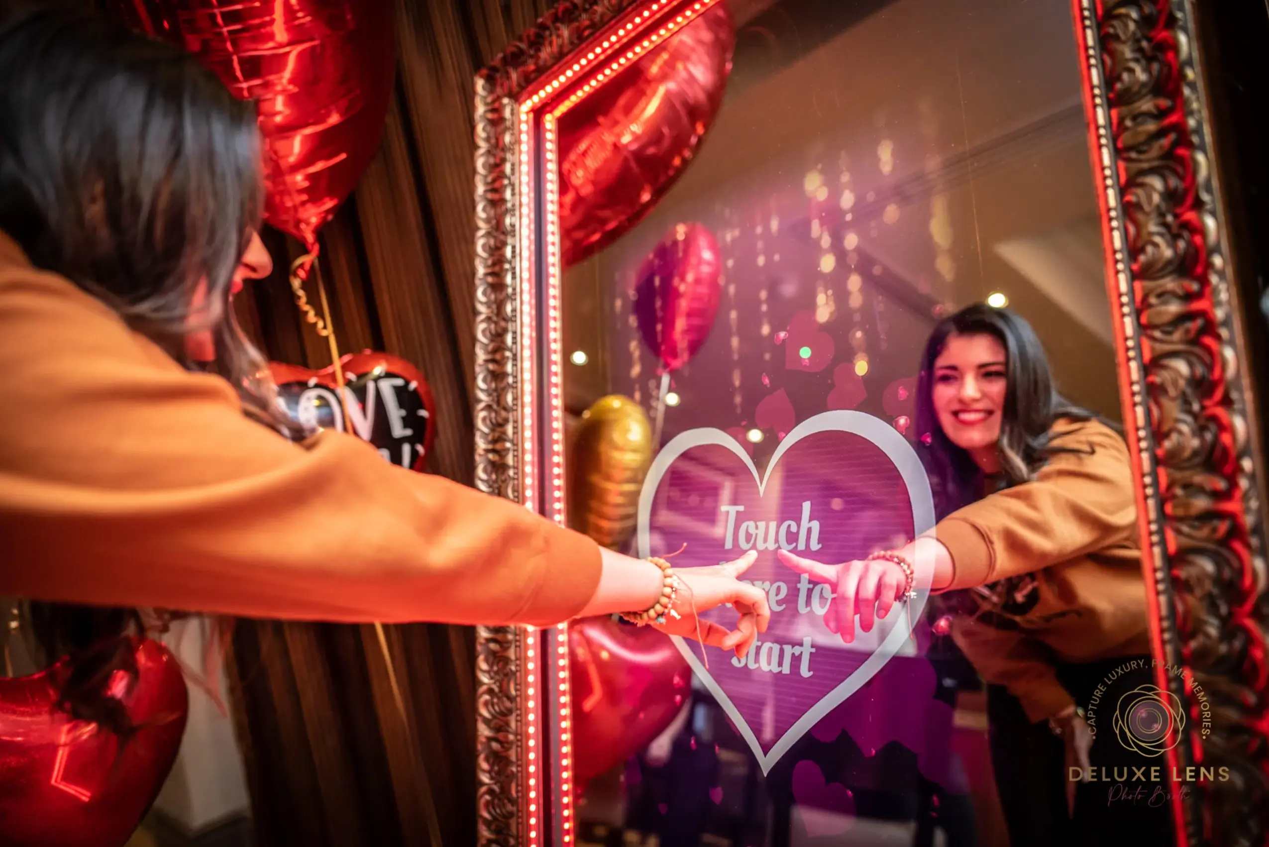 Woman looking at her reflection in a mirror with heart-shaped decoration, surrounded by red curtains and balloons, celebrating a special occasion.