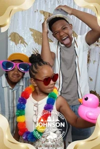 Three children wearing colorful accessories and sunglasses, smiling and playing with a pink rubber duck, indoors with a decorative background.