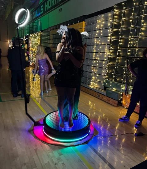 Two women stand on a colorful illuminated platform at a party, with festive lights and people in the background.
