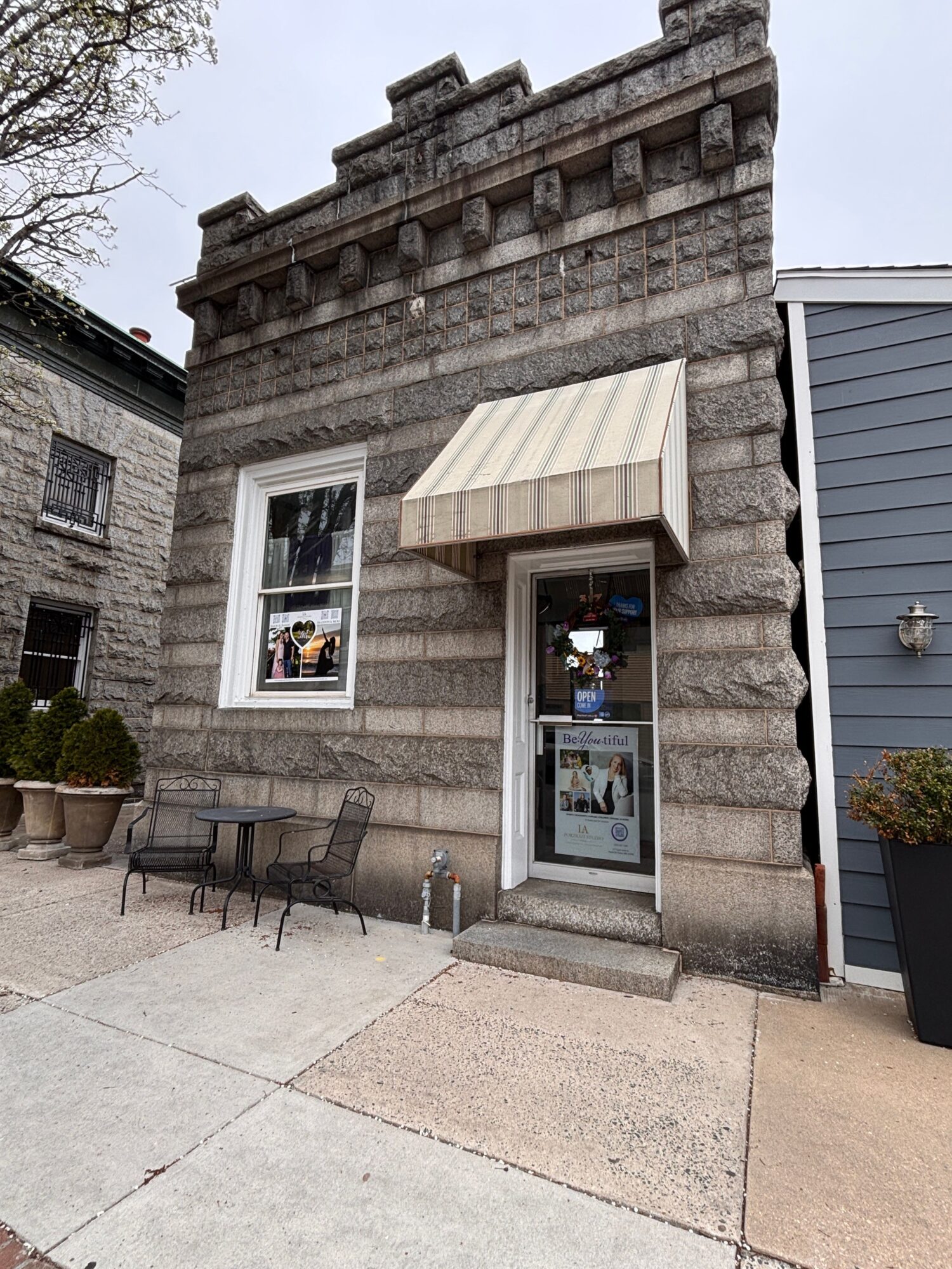 Small storefront with window display, awning, and outdoor bench, on a sidewalk with a stone wall and building in background.